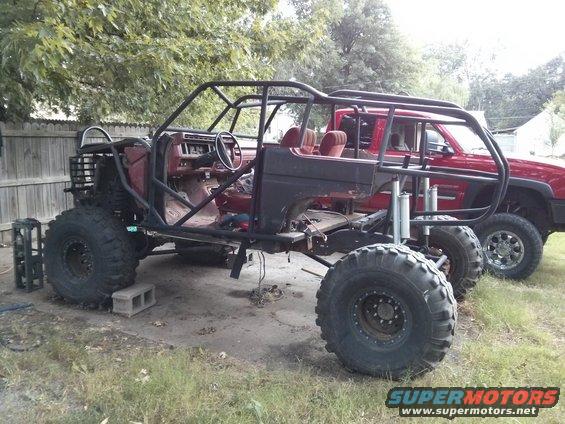 Modified off-road vehicle with roll cage, large tires, and red interior, parked next to a red pickup truck