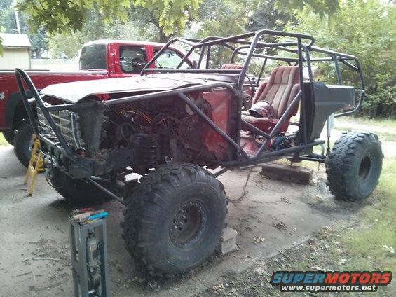 Custom buggy with roll cage, large tires, and exposed engine bay. Red pickup truck in background