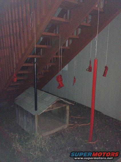 Red painted parts hanging from strings under wooden stairs next to a dog house