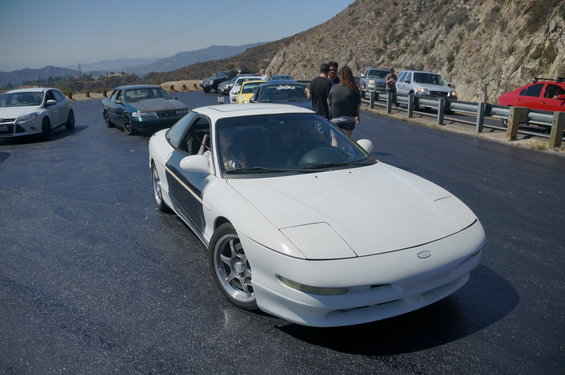 White coupe with a black stripe on the side, parked on a road with other cars. The car has pop-up headlights and aftermarket wheels
