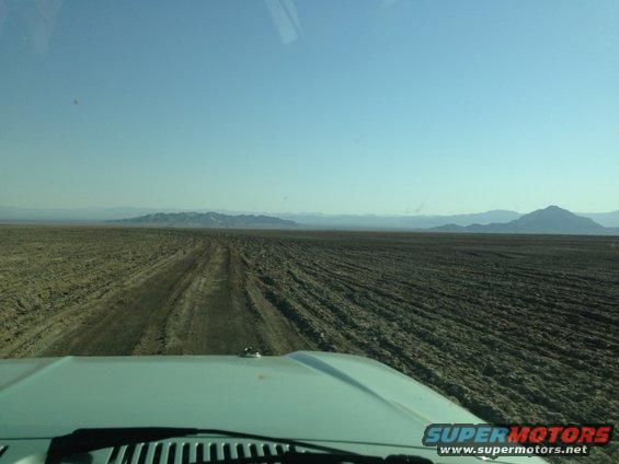 Light blue vehicle hood and windshield wipers in foreground, driving on a dirt track through a dry, barren landscape under a clear blue sky