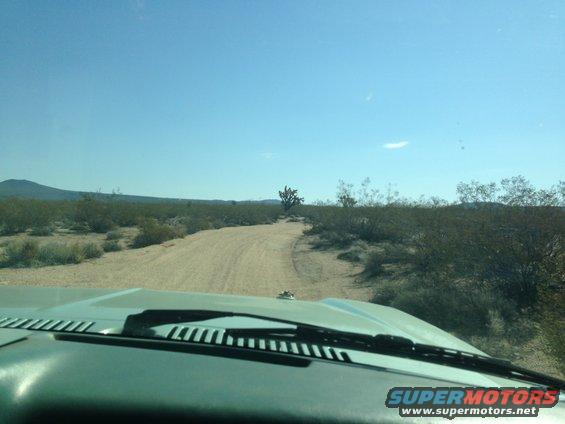 View from inside a vehicle looking out at a dirt road winding through a desert landscape with scrub brush and a distant mountain