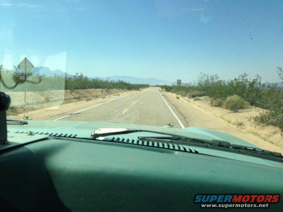 Teal dashboard and windshield showing a straight desert road with scrub brush and mountains in the distance