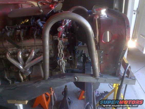 Red vehicle body with V8 engine and chrome exhaust headers, undergoing roll cage installation