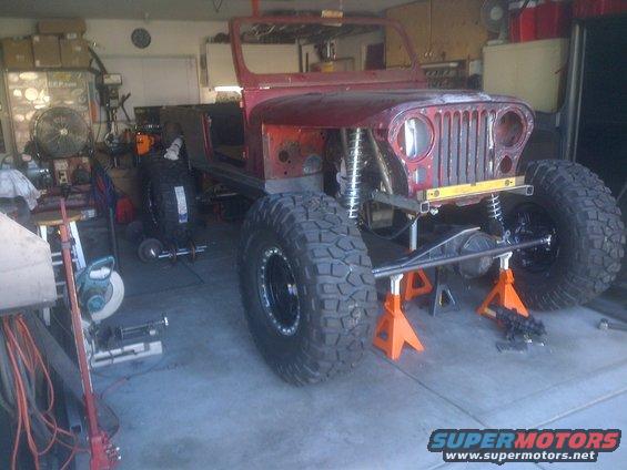 Red Jeep CJ undergoing modification, front view, raised on jack stands with large off-road tires