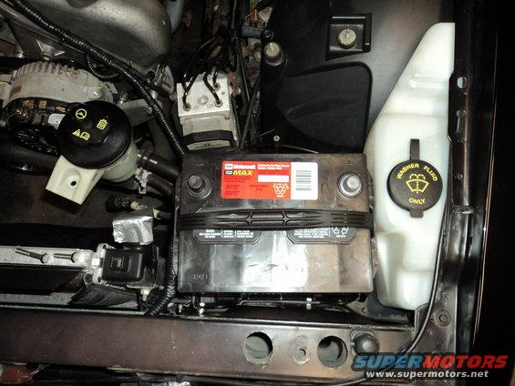Car engine bay showing a black battery with a red label and a white washer fluid reservoir