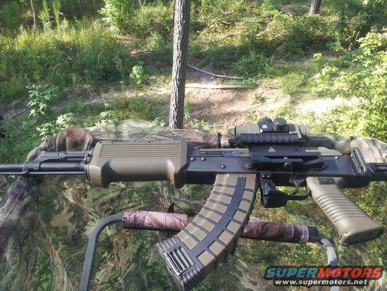 Assault rifle with scope and extended magazine, resting on camouflage material in a wooded area