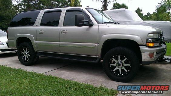 Silver SUV, 3/4 front view, with aftermarket wheels and tow mirrors. A white car is partially visible to the left, and a covered vehicle is behind