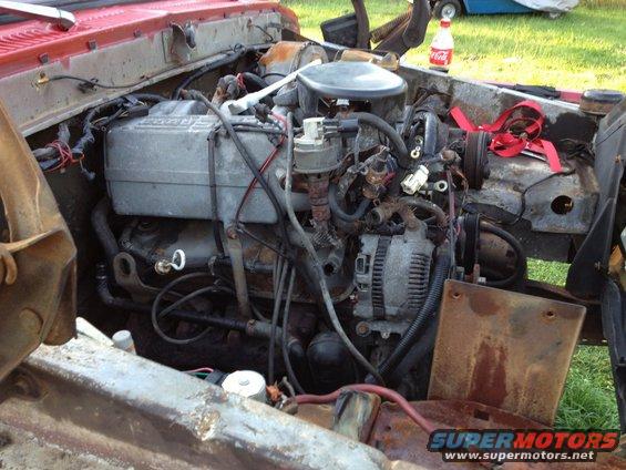 Engine bay of a red vehicle with a V8 engine, alternator, and various hoses and wires