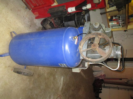 Blue air compressor with a large pulley wheel and metal tank, resting on a concrete floor