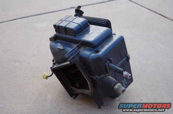 Black plastic automotive part with yellow connector and metal fittings, resting on concrete
