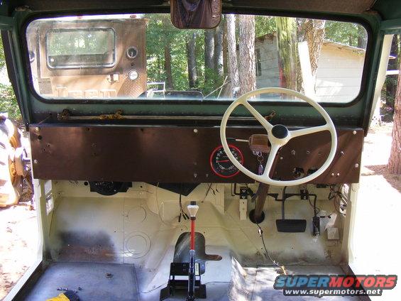 Interior view of a Jeep CJ with a white steering wheel and a red-faced gauge. Another vehicle is visible through the windshield