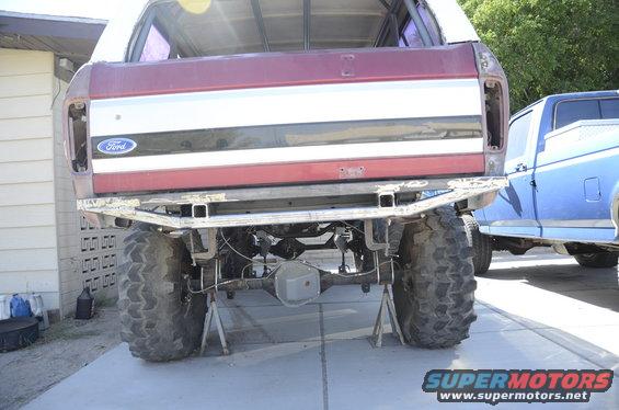 Rear view of a maroon Ford Bronco on jack stands, showing its differential and large tires. A blue pickup truck is parked nearby