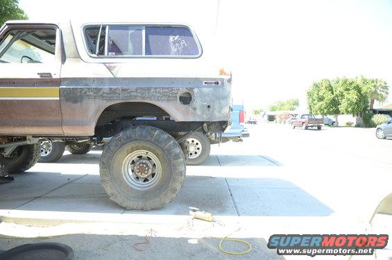 Side view of a lifted 1978 Ford Bronco with large tires, showing rust and missing rear bumper. Another vehicle is parked behind it