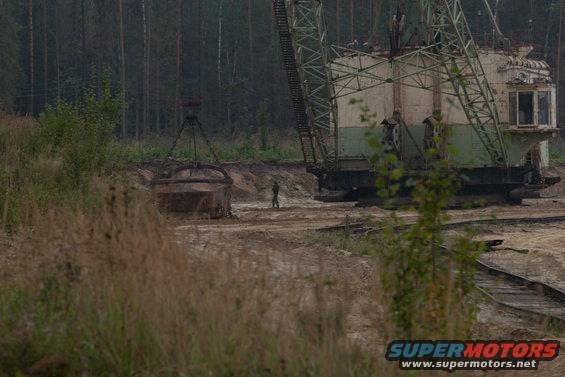 Giant excavator machine with a dragline bucket hanging nearby, a person stands between them in a dirt quarry