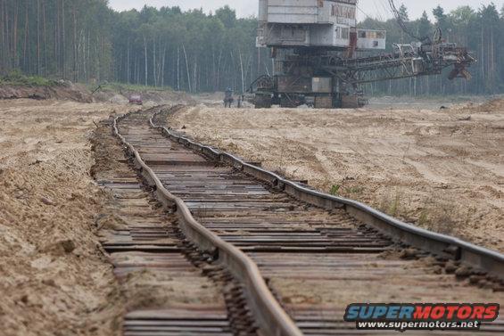 Railroad tracks curve through a sandy area towards a large excavator; a red vehicle is visible in the distance
