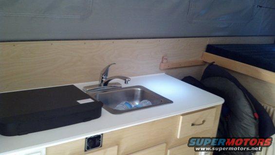 Camper interior showing a sink, faucet, black stove cover, and a bed in the background
