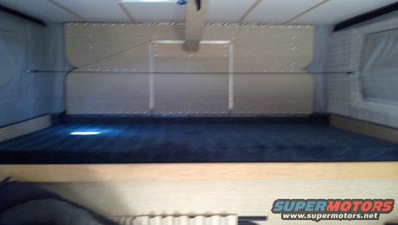 Interior of a camper with a dark blue mattress on a wooden platform. Walls are light wood paneling with metal accents