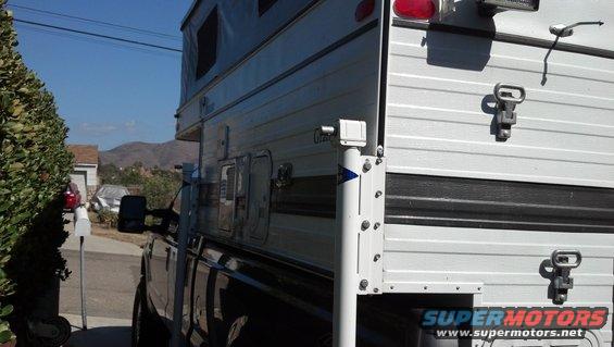 Truck camper with white siding and black trim, mounted on a dark truck. Visible are latches, a side mirror, and a pop-up roof section