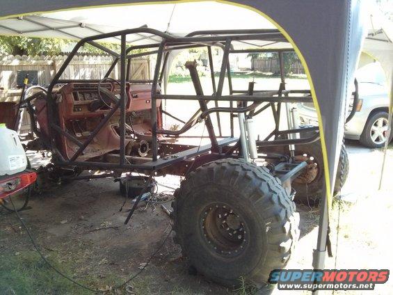 Off-road vehicle chassis with roll cage, red interior, large tires, and a white SUV in the background