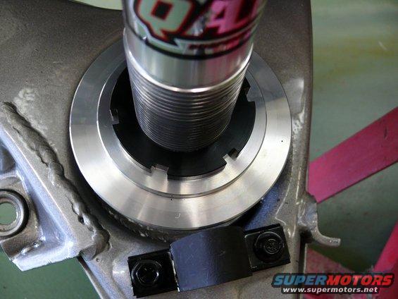 Close-up of a shock absorber assembly with a threaded adjustment collar and a black rubber strap secured by two bolts