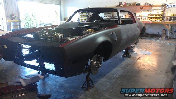 Unfinished car body, likely a 1970s Pontiac Firebird, on jack stands with engine exposed