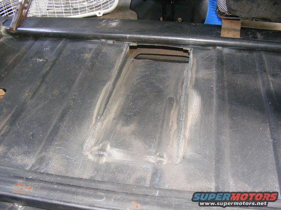 Black metal floor pan with a rectangular opening and visible weld repairs