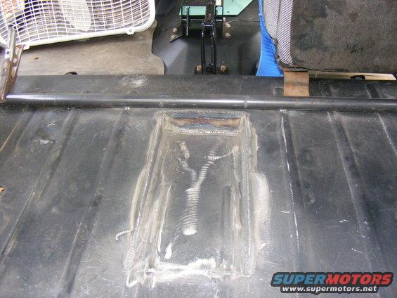 Close-up of a vehicle's metal floor with a welded repair patch. A seat and a fan are visible in the background