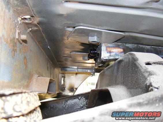 Close-up of vehicle frame with bolted metal plates and suspension parts. Some rust visible
