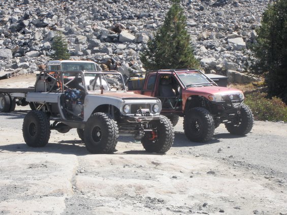 Two heavily modified off-road vehicles, one light-colored and one red, parked on a dirt road with rocky terrain in the background