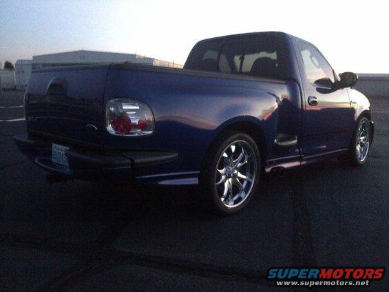 Blue pickup truck with chrome wheels, rear view. The vehicle has custom side skirts and tinted windows