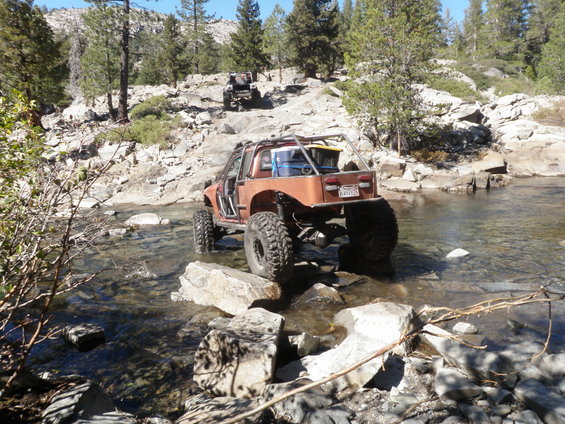 Orange off-road vehicle with large tires crossing a rocky stream, another off-road vehicle in background