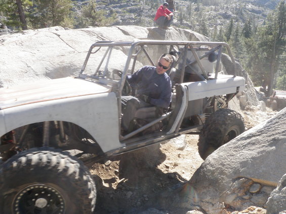 White off-road buggy with roll cage navigating rocky terrain, driver wearing sunglasses