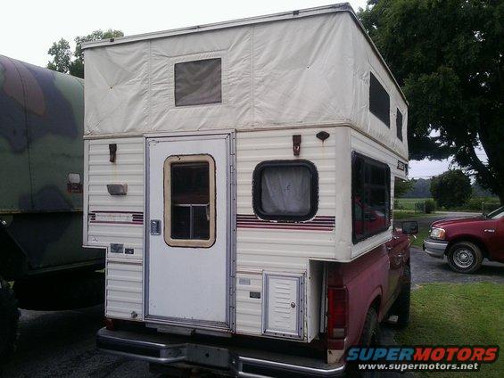 Rear view of a red pickup truck with a white pop-up camper shell. Another vehicle is parked to the right