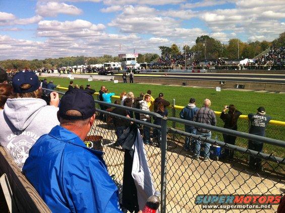 Crowd watching a drag race from bleachers, with a track and trees in the background