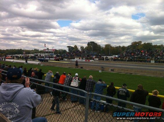 Spectators watch a drag race from the stands. Cars are on the track, and crowds fill the bleachers under a cloudy sky