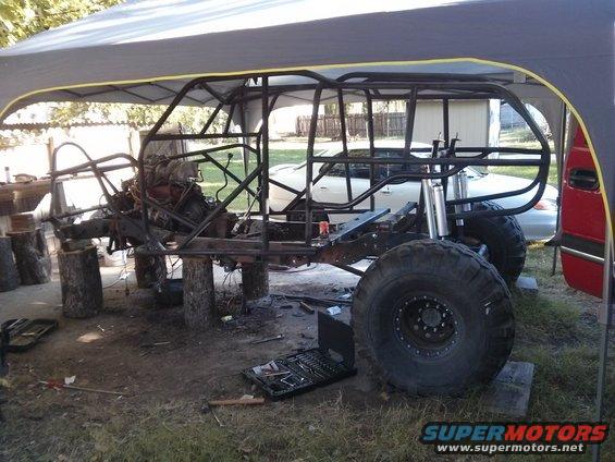 Off-road vehicle chassis with engine and suspension components, supported by logs. Tools are on the ground