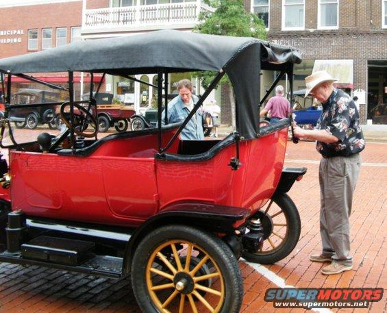 1909 Ford Model T touring car, red with black top, parked on brick street with two men nearby
