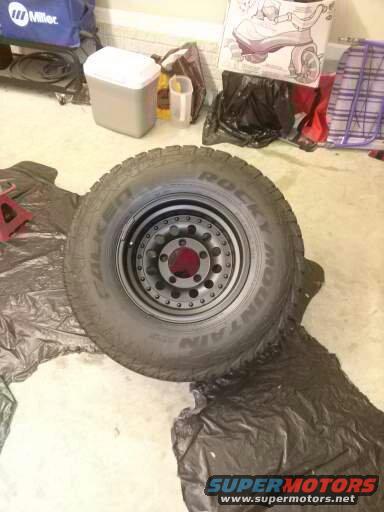 Tire and wheel assembly on black plastic sheeting, with Supermotors.net logo at bottom right