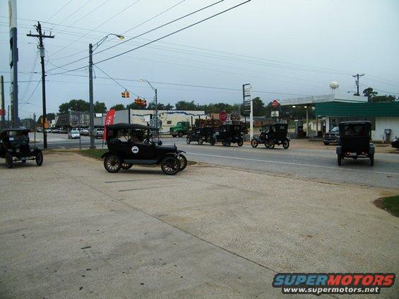 Several 1909 Ford Model T cars driving in a group past a gas station