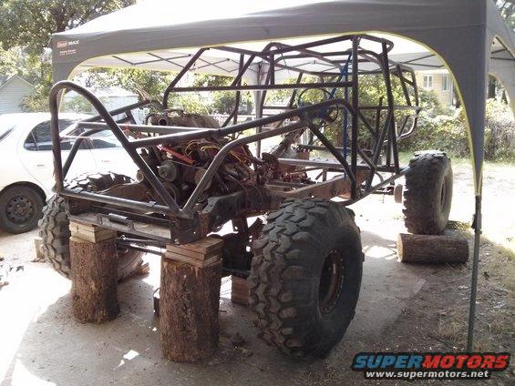 Buggy frame on logs, engine exposed, big tires, white car in background