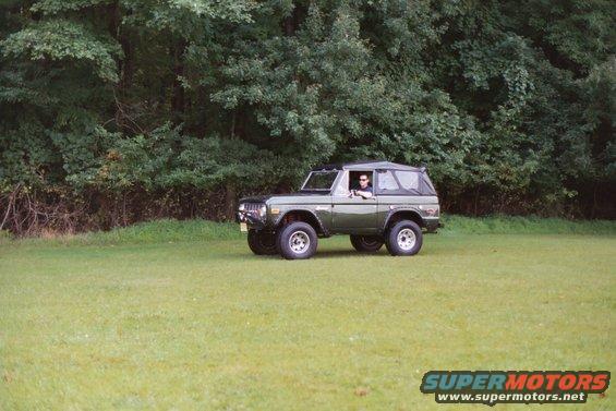 Green 1976 Ford Bronco with a person driving, parked on a grassy field with trees in the background