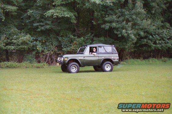 Green 1976 Ford Bronco parked on grass with trees in background. Driver visible