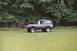 1976 Ford Bronco - A driver is behind the wheel of a green Ford Bronco as it sits on a grassy field with trees behind it.
