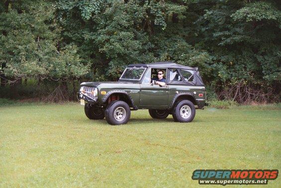 Green 1976 Ford Bronco with a black soft top and a man driving, parked on a grassy field with trees in the background