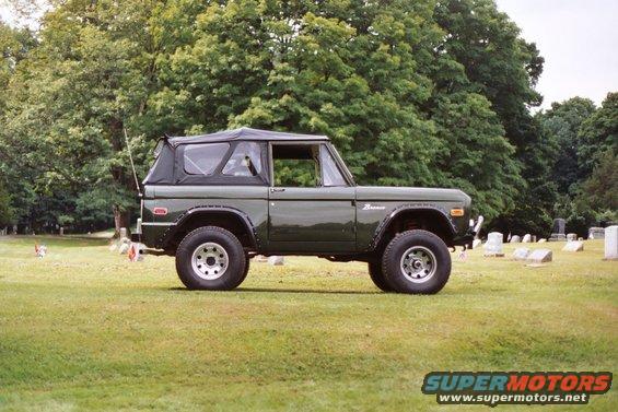 Green Ford Bronco parked in a grassy area with trees and gravestones in the background