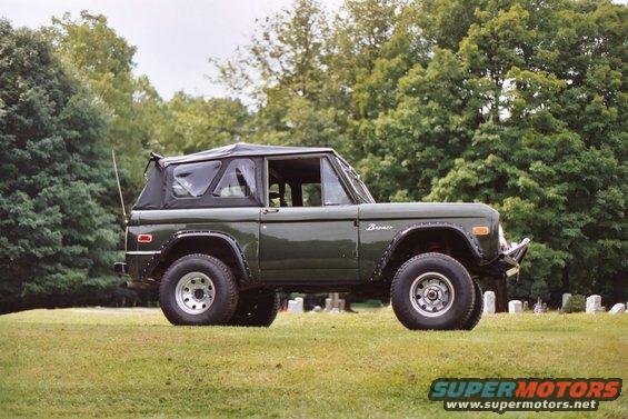Green 1976 Ford Bronco with a black soft top, parked on grass with trees and gravestones in the background