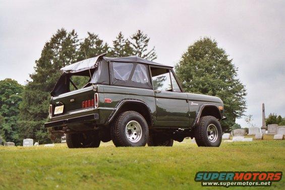 Dark green 1976 Ford Bronco with soft top, parked on grass with trees and gravestones in background