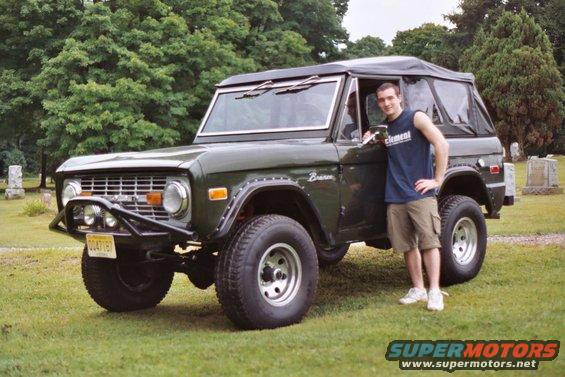 Dark green 1976 Ford Bronco with a soft top and aftermarket bumper, parked on grass with a man standing beside it