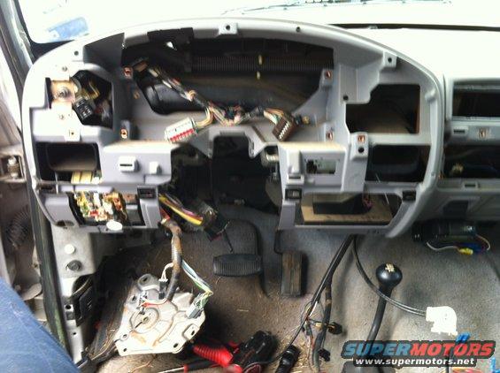 Vehicle dashboard with panels removed, showing wiring and components. Tools are visible on the floor